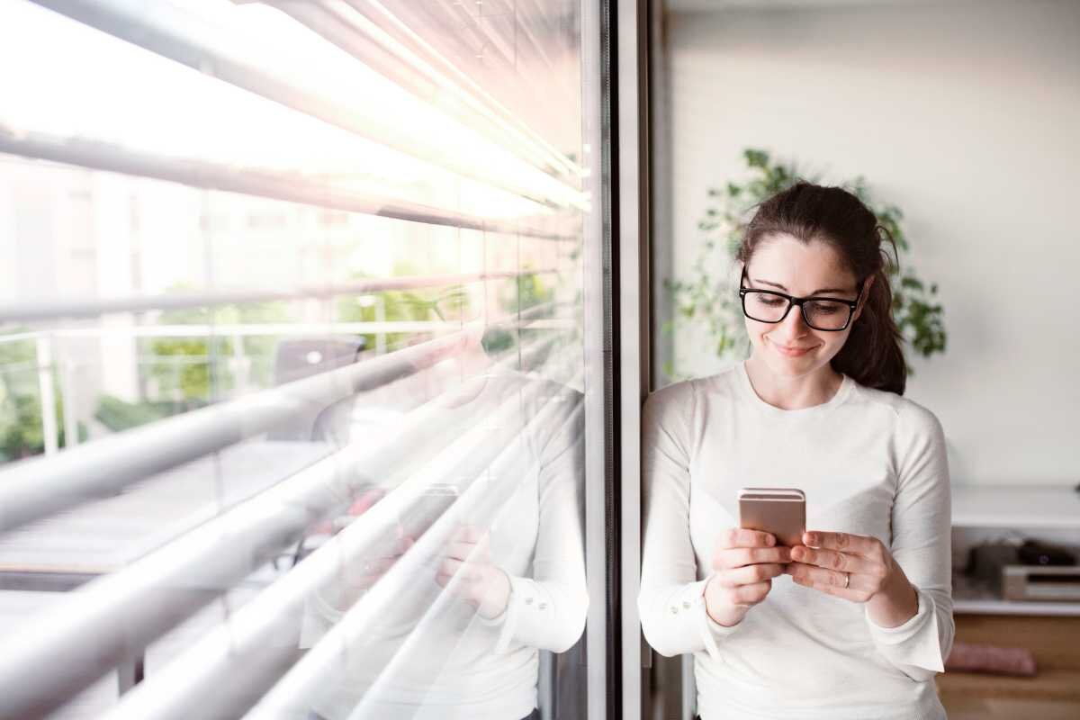 Fenster Aussenjalousien mit Smartphone-Seteuerung Frau mit Smartphone lehnt am Fenster mit Jalousien